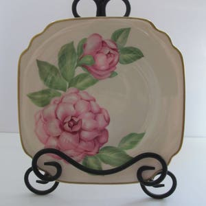 May include: A square, cream-colored plate with a gold rim features a pink floral design. The plate is on a black metal stand.