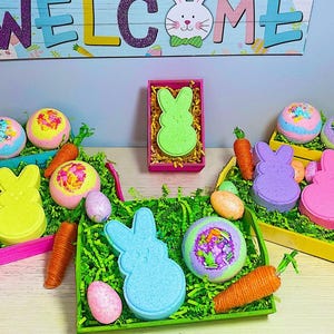 Könnte beinhalten: Sortiment an bunten Badebomben und Hasenförmigen Badebomben in verschiedenen Farben, darunter rosa, gelb, grün und blau. Die Badebomben sind in Körben mit grünem Gras ausgelegt und mit Ostereiern dekoriert.