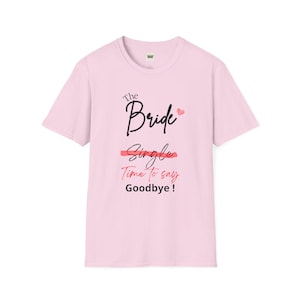 Op de afbeelding: Roze T-shirt met de tekst "The Bride" in zwarte schrijfletters met een klein roze hartje. De tekst "Single" is doorgestreept met een rode lijn en de tekst "Time to say Goodbye!" is in zwart.