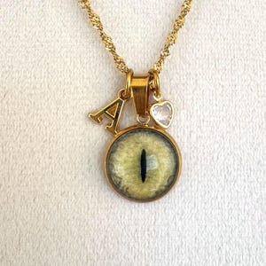 Könnte beinhalten: Goldfarbene Halskette mit einem Anhänger mit einem grünen und schwarzen Augendesign. Die Halskette enthält einen goldenen Buchstaben "A" Anhänger und einen kleinen herzförmigen Anhänger. Die Kette hat ein gedrehtes Seildesign.