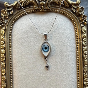 May include: A silver-toned necklace featuring an eye-shaped pendant with a realistic blue iris and white sclera, set in a metallic frame. A small, decorative silver-toned metal charm with an intricate filigree pattern hangs below the eye pendant.