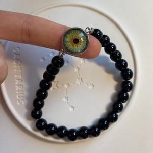 May include: A stretch bracelet featuring smooth, round, black beads and a central circular charm displaying a realistic, detailed green human eye with a black pupil and golden-brown iris accents, set in a polished silver-toned metal frame.