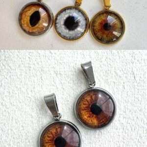 Pode incluir: Colares com pingentes de olhos com argolas em aço inoxidável e douradas. Os pingentes apresentam desenhos detalhados de olhos em tons de castanho, branco e roxo. As joias são exibidas em uma superfície branca texturizada.