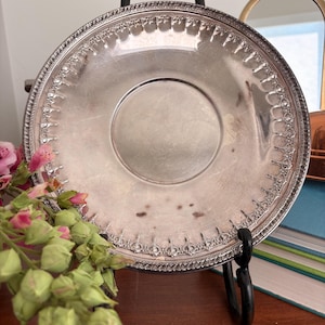 May include: A round silver-toned metal saucer featuring a decorative embossed border with repeating arched patterns. The center of the saucer is smooth with visible signs of tarnish and light surface wear, reflecting a metallic sheen throughout.