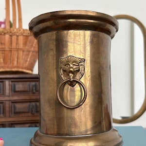 Puede incluir: Un recipiente cilíndrico de latón con un emblema de cabeza de león y un tirador de anillo circular en la parte frontal. La superficie metálica muestra una pátina cálida y envejecida, con un borde ancho y acampanado y una base circular.