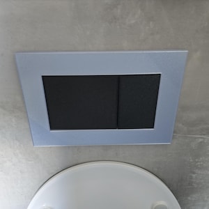 Pode incluir: Uma placa de descarga retangular azul claro com dois botões retangulares pretos. A placa está montada numa parede cinza claro, com um objeto semicircular branco visível abaixo.