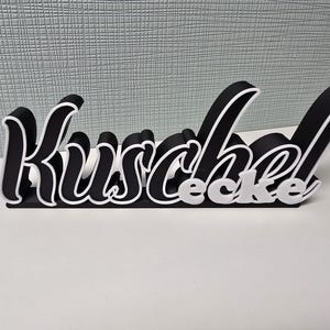 Könnte beinhalten: Ein schwarz-weißes Dekoschild mit der Aufschrift "Kuschelecke" in einer stilisierten Schriftart. Die Buchstaben sind schwarz mit weißen Konturen, und das Wort "ecke" ist weiß. Das Schild steht auf einer weißen Oberfläche.