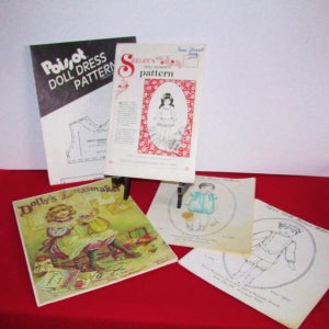 May include: A collection of vintage doll dress patterns and books. The patterns include "Polisot Doll Dress Pattern" and "Seeley's Doll Maker's Pattern." The books feature illustrations of dolls and dressmaking instructions. The items are on a red surface.