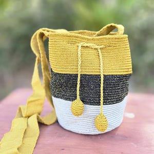 Op de afbeelding: Een handgemaakte gehaakte emmer tas in mosterdgeel, zwart en wit. De tas heeft een trekkoordsluiting met gele kwastjes en een lange, verstelbare riem. De tas is ongeveer 25 cm hoog.