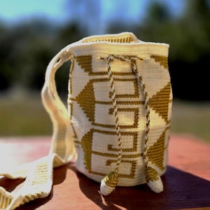 May include: Cream-colored, hand-crocheted shoulder bag with a geometric pattern in gold. The bag features a long strap and drawstring closure with tassels. The bag is approximately 10 inches tall and 8 inches wide.
