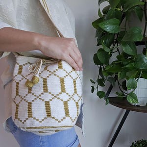 May include: A woven bucket bag featuring a white base with a repeating geometric pattern in olive green. The bag includes a matching olive green drawstring closure with two spherical tassels at the ends and a long, wide woven shoulder strap.