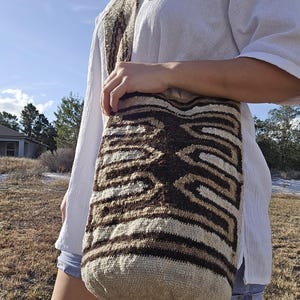 May include: A woven shoulder bag with a geometric pattern in shades of brown, beige, and cream. The bag has a matching strap and is worn over the shoulder. The bag is made of a textured material.