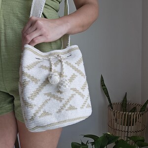 May include: White and gold crochet bucket bag with a drawstring closure and two decorative pom-poms. The bag features a geometric pattern and a matching shoulder strap. The bag is held by a person wearing a green romper.