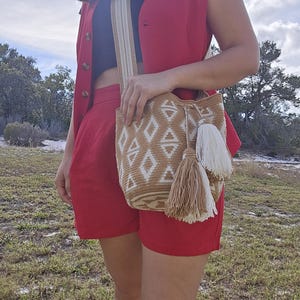 May include: A tan and white woven shoulder bag with a geometric diamond pattern and two tassels. The bag has a wide strap and is worn with a red vest and shorts. The outfit is completed with a black top.