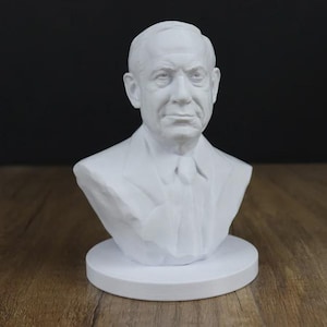 May include: A white bust sculpture of a man with short hair, a suit jacket, and a tie. The sculpture is on a round, white base. The background is dark, and the sculpture is on a wooden surface.