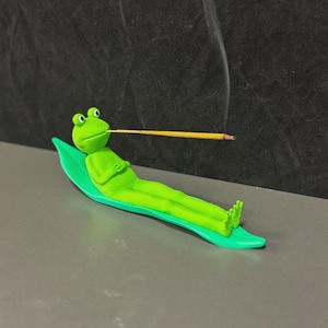 Pode incluir: Um porta-incenso de sapo verde. O sapo está reclinado sobre uma base verde em forma de folha, segurando um incenso na boca. A fumaça sobe do incenso aceso, criando um ambiente relaxante.