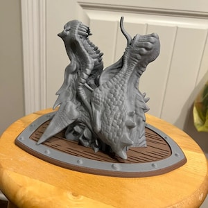 Może przedstawiać: Sztuka głowy smoka z dwoma głowami, wydrukowana w 3D w kolorze szarym, zamontowana na brązowej drewnianej tarczy z srebrną metalową obwódką.