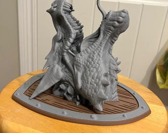 May include: A 3D printed gray dragon head sculpture with two heads, mounted on a brown wooden shield with a silver metal trim.