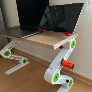 May include: Adjustable laptop and tablet stand. The stand is white with green and orange accents. A laptop and tablet are placed on the light brown surface. The stand is designed to elevate devices for ergonomic use.