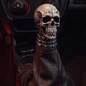 Może przedstawiać: Gałka zmiany biegów w kształcie czaszki z kolczastym kołnierzem, zamontowana na czarnej skórzanej osłonie dźwigni zmiany biegów z czerwonymi szwami. Czaszka jest szara i szczegółowa, dodając unikalnej, gotyckiej estetyki do wnętrza samochodu.