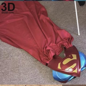 May include: A red Superman cape with a burgundy collar and the iconic gold and burgundy emblem. The cape is draped over a blue and burgundy helmet. The text "3D Do3d.com" is visible in the upper left corner.