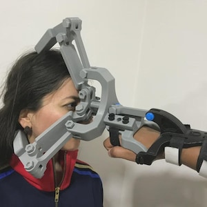 Può includere: Un dispositivo meccanico grigio è tenuto davanti al viso di una persona e attaccato al suo polso. Il dispositivo ha più bracci articolati e un tutore per il polso nero con cinturini bianchi. La persona indossa una giacca blu, rossa e bianca.