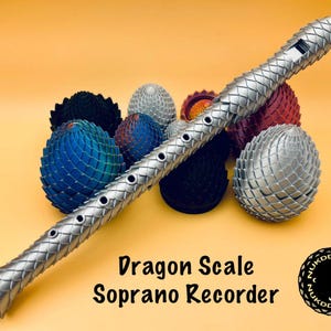 May include: A silver soprano recorder with a dragon scale design, alongside a collection of colourful, dragon-egg-shaped sculptures. The text "Dragon Scale Soprano Recorder" is visible. The background is a solid orange colour.