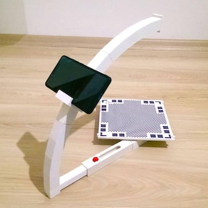 May include: A white, curved stand with a black tablet secured at the top. A checkered pattern board is positioned below. The stand has a red button and is designed to hold a tablet for various uses.