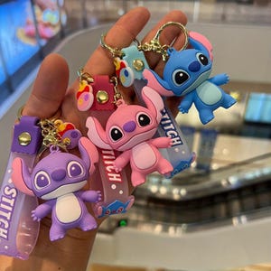 May include: A selection of Stitch character keyrings in blue, pink, and purple. Each keyring features a Stitch figure with a clear strap that says "Stitch". The keyrings are attached to gold-coloured metal rings.
