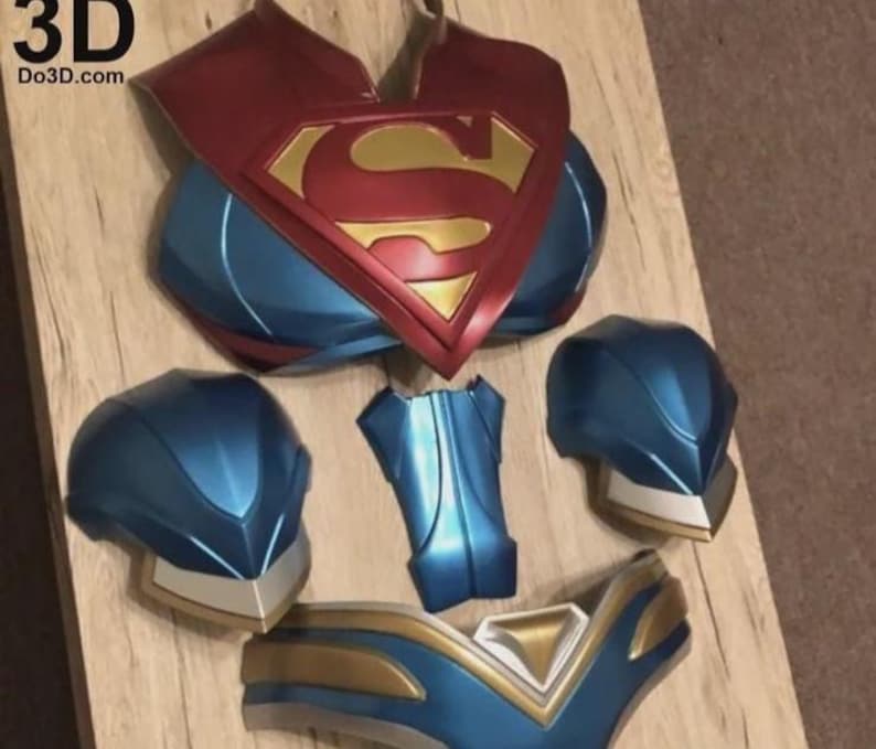 May include: A disassembled Superman costume, with a red and gold chest emblem, blue and gold shoulder pieces, and blue helmet components. The text "3D Do3D.com" is visible. The pieces are arranged on a wooden surface.