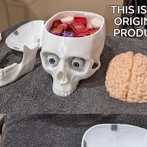 May include: A white skull-shaped container with a removable lid, jawbone, and a separate brain model. The skull's eye sockets contain blue eyeballs. The container is filled with wrapped candies. Text on the image reads "THIS IS AN ORIGINAL PRODUCT."