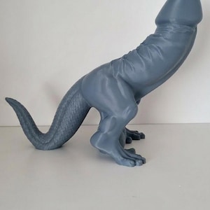 May include: A grey sculpture of a dinosaur-like creature with a phallic form. The sculpture has a long tail, two legs, and is made of a smooth material. It is positioned on a white surface.