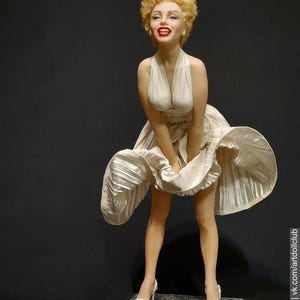 Peut inclure: Poupée de Marilyn Monroe dans une robe blanche à col licou, debout sur une surface noire. La robe est plissée et flottante, avec la jupe relevée. La poupée a les cheveux blonds, du rouge à lèvres rouge et des talons hauts blancs.