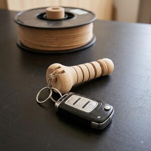 Può includere: Un portachiavi in legno di colore chiaro con un design cilindrico scanalato, fissato a un telecomando per auto elettronico nero e argento tramite un anello metallico. L'impugnatura in legno presenta un'estremità arrotondata e una tacca.