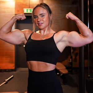 Op de afbeelding: Een vrouw in een zwarte sportbeha en leggings poseert met haar armen gebogen, waardoor haar gespierde lichaam wordt getoond. Ze bevindt zich in een sportschool met gewichten en apparatuur op de achtergrond.