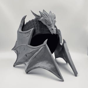 May include: A gray fantasy helmet with a dragon design. The helmet features a dragon head crest, wing-shaped side pieces, and a scale-like texture on the top. The helmet is a cosplay or costume accessory.