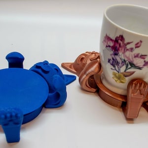 May include: A blue and a copper-colored 3D-printed mug holder in the shape of a cartoon character. The blue holder is lying on its back. The copper holder is holding a white mug with a floral design.