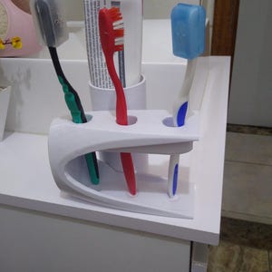 May include: A white plastic toothbrush holder containing three toothbrushes and a tube of toothpaste. The toothbrushes are red, blue, and green, with protective covers. The toothpaste tube is white with red and blue text. The holder is on a white surface.