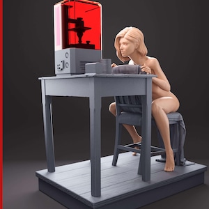 Könnte beinhalten: Eine 3D-gedruckte Figur einer Frau, die auf einem Stuhl an einem Tisch sitzt und eine rote 3D-Druckmaschine betrachtet. Die Figur ist aus hellgrauem Material gefertigt und auf einem Holzsockel platziert.