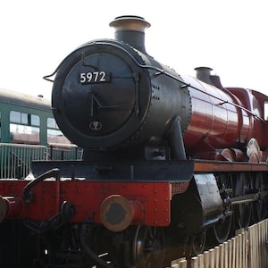 May include: A vintage steam locomotive, painted in black and maroon, with the number 5972 displayed on its front. The train is parked next to a green passenger car. The locomotive has a red front and is positioned on tracks.
