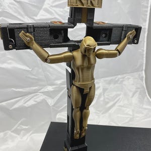 Può includere: Un crocifisso stampato in 3D con una figura articolata color oro su una croce nera e oro. La croce ha le lettere INRI in alto. Le braccia della figura sono tese e il design ha un'estetica meccanica.