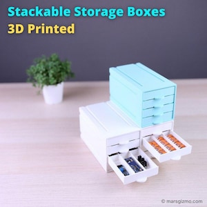 May include: Stackable storage boxes, 3D printed, in white and light blue. The boxes have pull-out drawers, some of which are open, revealing small electronic components. The text "Stackable Storage Boxes 3D Printed" is at the top.