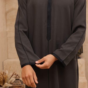 May include: Dark gray thobe with black trim and detailing. The long-sleeved garment features a mandarin collar and a decorative black strip down the front. The sleeves are cuffed with black trim. The thobe is a traditional Middle Eastern garment.