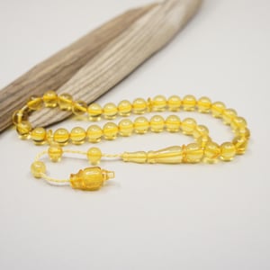 Puede incluir: Un collar de cuentas de oración de color ámbar con borla y pieza final decorativa. Las cuentas son redondas y están ensartadas en un cordón de color claro. Un trozo de madera flotante descansa en el fondo.