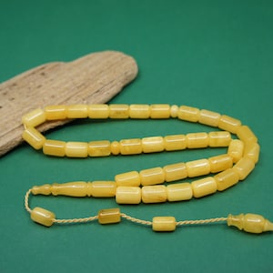 Peut inclure: Un chapelet composé de perles cylindriques lisses de couleur jaune pâle. Les perles sont reliées par un cordon fin de couleur claire, se terminant par un élément décoratif avec des perles plus petites et une pièce terminale sculptée.
