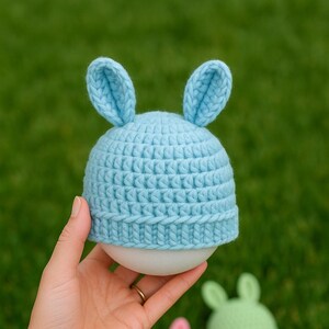 May include: A light blue crocheted bunny hat with upright ears, held in a hand. Three other bunny hats in pink, green, and yellow are in the background, all on white spheres. The hats are on a green grass background.