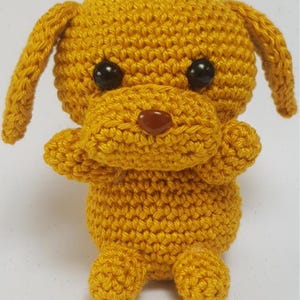 Könnte beinhalten: Ein kleiner, gehäkelter gelber Welpe mit schwarzen Augen und einer braunen Nase. Der Welpe sitzt mit gekreuzten Beinen.