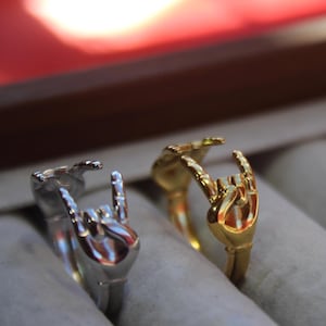 May include: Two rings with a hand gesture design. One ring is gold and the other is silver. The rings are displayed on a white surface.