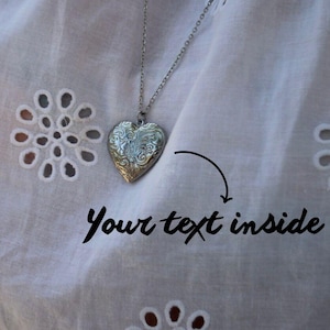 Puede incluir: Un collar con un colgante plateado en forma de corazón adornado, suspendido de una fina cadena, sobre una tela blanca con ojales. El texto "Your text inside" está escrito debajo.