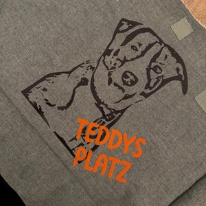 Könnte beinhalten: Eine graue Canvas-Tragetasche mit einer schwarzen Silhouette eines Hundes und dem Text "Teddy's Platz" in orangefarbener Stickerei.